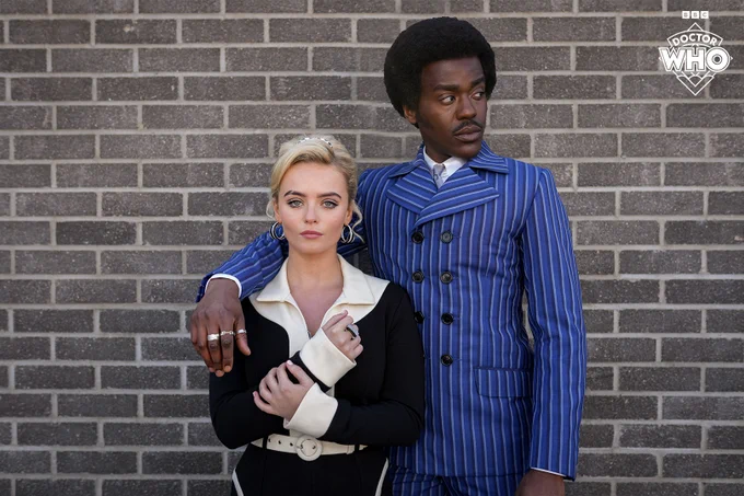 Ncuti Gatwa and Millie Gibson as the Doctor and Ruby Sunday, dressed in 1960s-style clothing, posing in front of a brick wall.