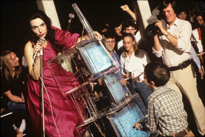 Cellist Charlotte Moorman plays Nam June Paik's TV Cello while a crowd watches and a man films her.