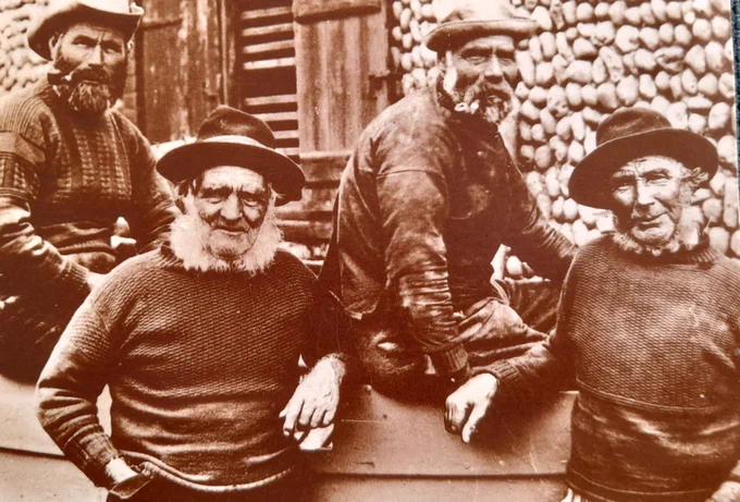 Sepia photo of four elderly fishermen, posing by and on an upturned boat. They look to be Victorian in era with lots white facial hair, battered hats and knitted jumpers. Two of them are smoking pipes.