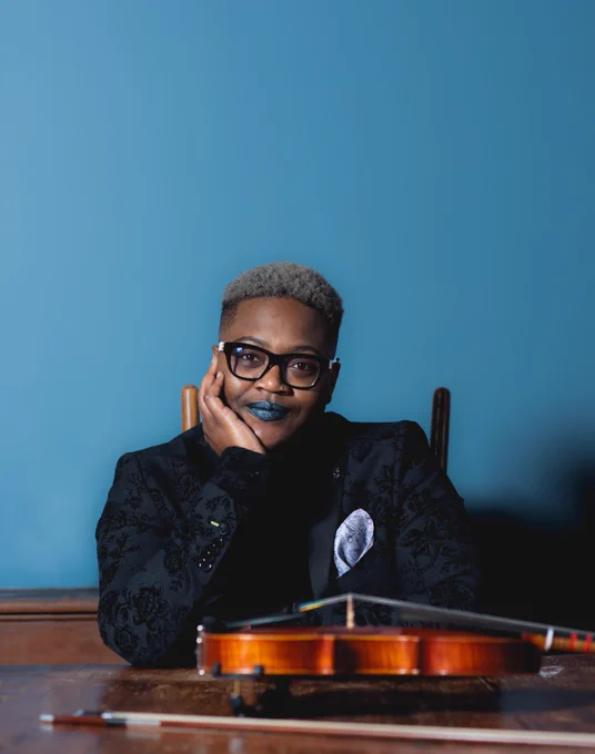 Headshot of a Black person seated with their right elbow on a wooden table with their right hand framing their face. They are wearing a black and cream glasses, a black suit with velvet black floral print, steel grey floral pocket square, gunmetal blue lipstick, and a soft smile. Laying flat in front of them is a stradivarius model violin and its bow. Photo credit Elliott Tilleczek