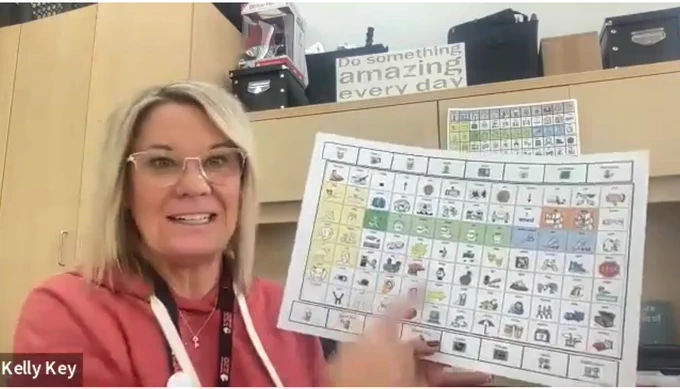 A woman sitting in an office area holding and pointing to a communication board with symbols with a sign behind her that reads "Do something amazing every day."