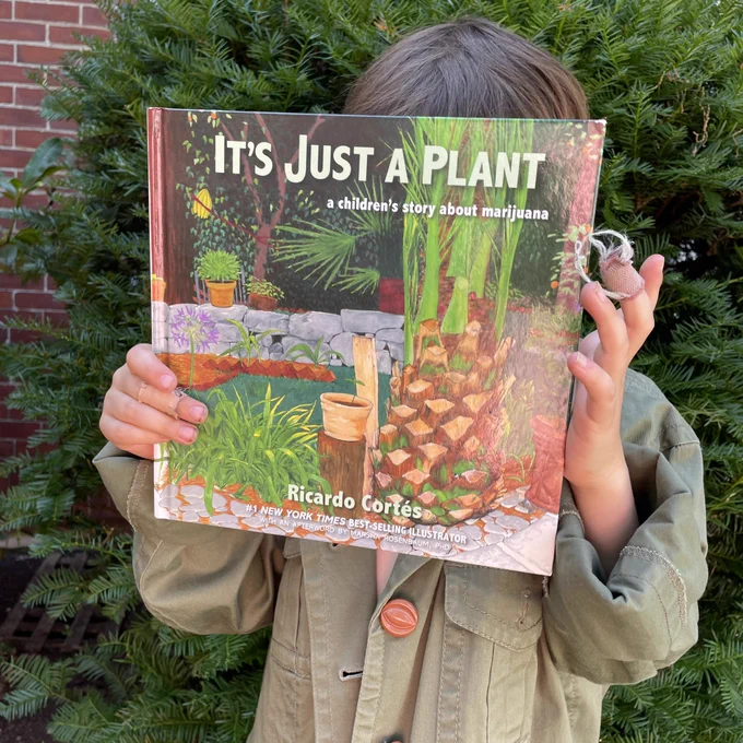 A photo of a child holding up a book. The book is “It’s Just a Plant - a children’s story about marijuana,” and has an illustration on the book cover of a small potted plant sitting on a log, amidst a garden with several more plants and stones around it.