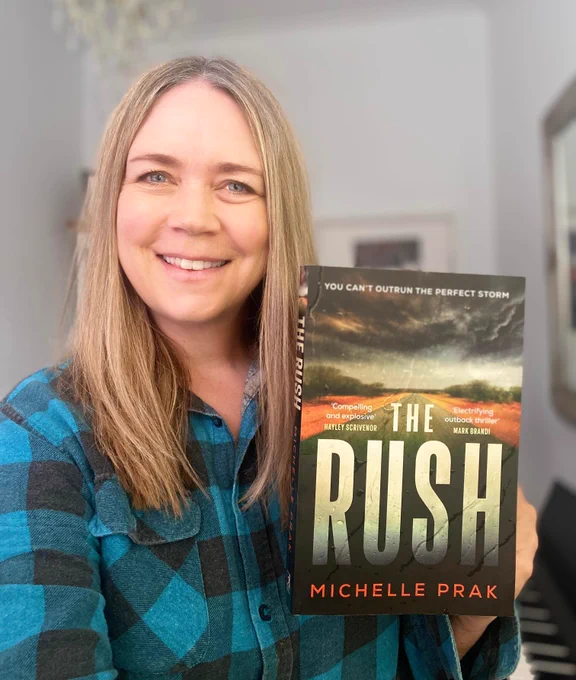 Michelle Prak holds up a copy of her novel, "The Rush". She's smiling, with long straight hair, and wearing a blue and black checked flannel shirt.