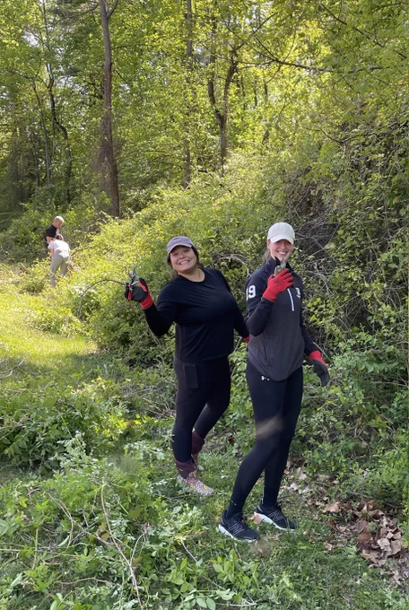 Two Burness staff smile at the camera during a volunteer service project. They are surrounded by green space. 