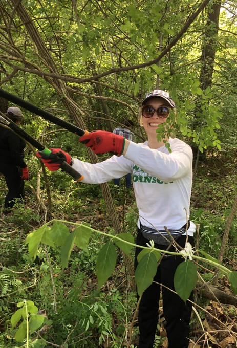 A Burness staff member uses trimmers to cut invasive plants near a small park in Bethesda. She's wearing sunglasses and a hat. 