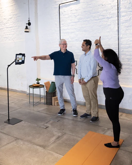 Tim is standing next to the founders of Prayoga. One founder is looking at an iPad on a stand and conducting yoga poses on an orange yoga mat.