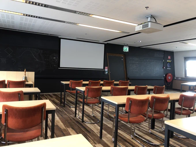 Photo of a large well lit room set up for a lecture or seminar with rows of tables and orange chairs facing the front of the room. There’s a project screen at the front of the room, and blackboards either side covered in writing. There’s a big bell on one of the tables at the front of the room.