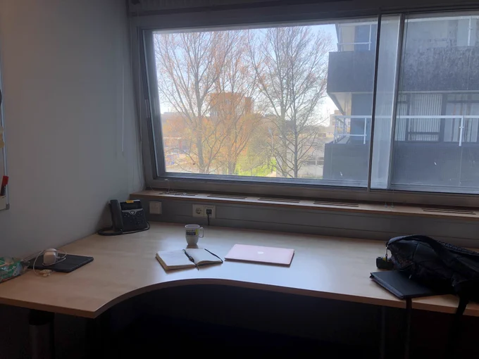 Photo of a desk in front of a window looking out onto a sunny day with some trees and university buildings outside. On the desk is a laptop and notebook, and a mug, as well as a backpack on the neighbouring desk.