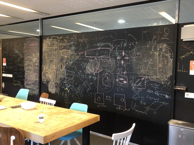 Photo of a brightly lit room with a wall covered with a blackboard, covered in overlapping writing and diagrams in brightly coloured chalk. In front is a large wooden table with lots of chairs and a few mugs and bowls of snacks.