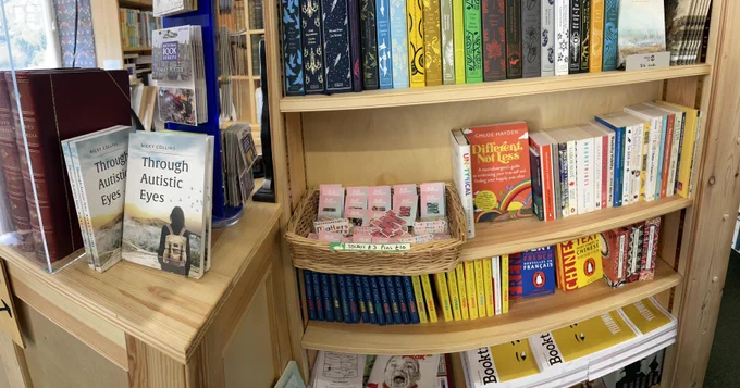 A pleasant looking bookshelf with a selection of books about Autism Spectrum Disorder. Also on the shelf are a collection of cute enamel pins and stickers about ASD and neurodivergence.