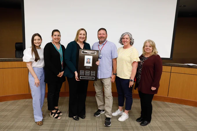 Six people smile while holding a plaque.