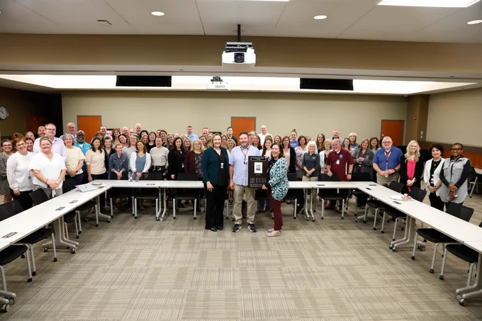 A large group of people smile for a man receiving a plaque.