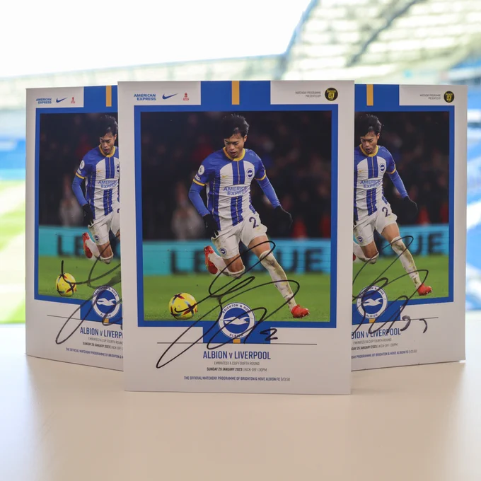 Three match programmes from Albion's game against Liverpool, with Mitoma's signature on the front. The arches of the Amex can be seen in the background.