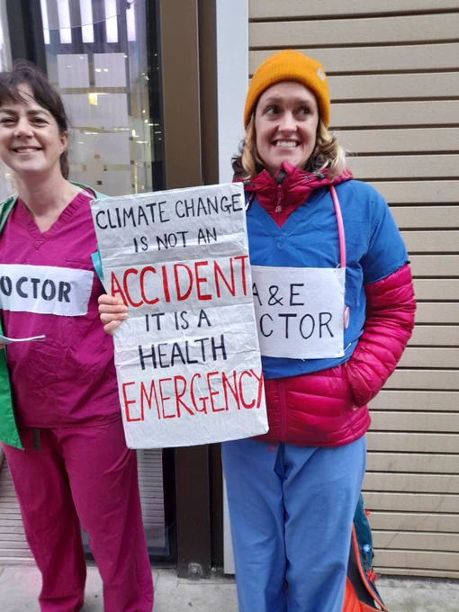 Two doctors wearing scrubs hold a sign that says „climate change is not an accident, it is a health emergency“