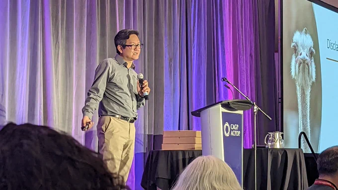 Vu Le speaking on stage with a picture of a slide of an angry ostrich in the background.