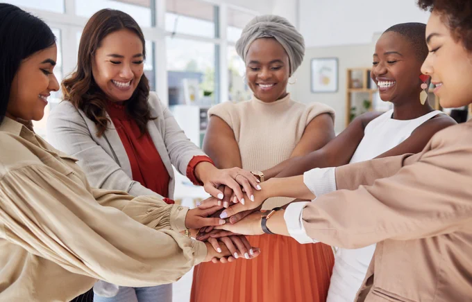 A group of smiling young women from the Global Majority stand in a tight group with their hands outstretched and overlapping in a gesture of solidarity