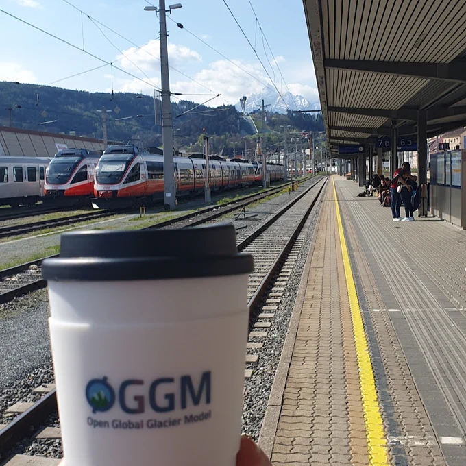 Foreground: hand holding a reusable coffee cup with the Open Global Glacier Model logo on it. Background: Innsbruck central train station and snowy mountains.