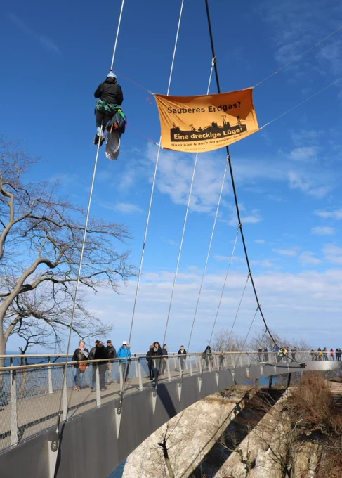Bild zeigt eine Stahlkonstruktion auf der Menschen laufen und den Ausblick auf das Meer genießen. An Stahlseilen ist eine Person in die Höhe geklettert und zeigt ein Banner mit der Aufschrift: "Sauberes Gas ist eine dreckige Lüge."