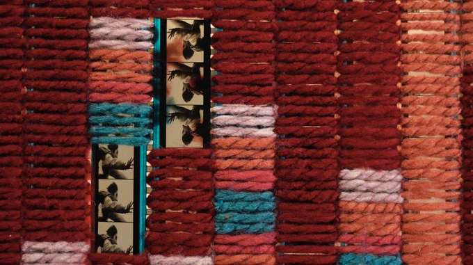 ocumentation of Tahir's works. The work is rows of yarn, predominantly in red, with hints of pink, orange, fuschia, and teal. The yarn is woven around sepia toned film.