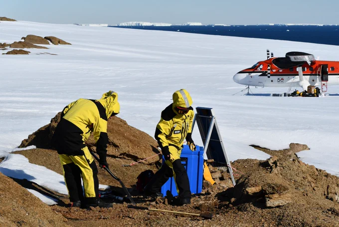 Two field scientists at work upgrading a seismic station on a rock outcrop, a red twin otter aircraft waits in the background, and icebergs are visible in the far distance.