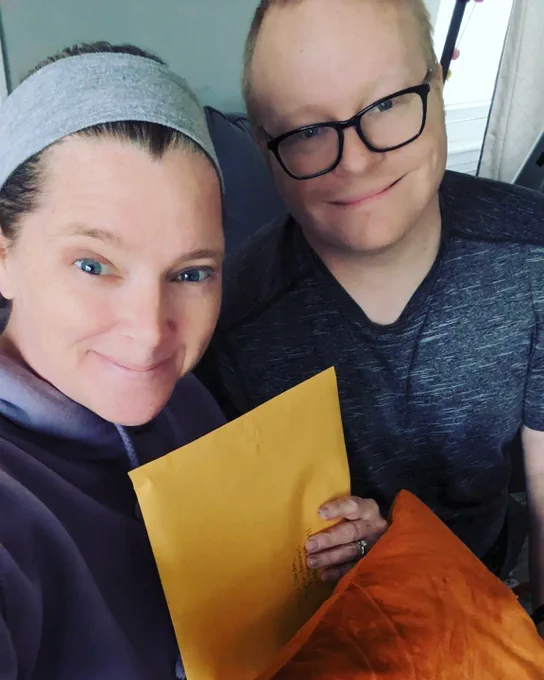 Katy and Trevor hold a vanilla envelope holding an orange pillow 
