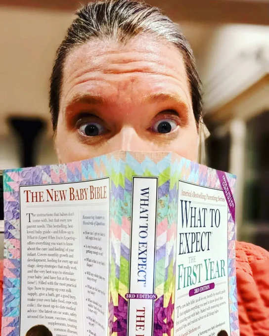 Katy holds the book: What To Expect In The First Year up against her face so you only see her shocked, slightly overwhelmed eyes 
