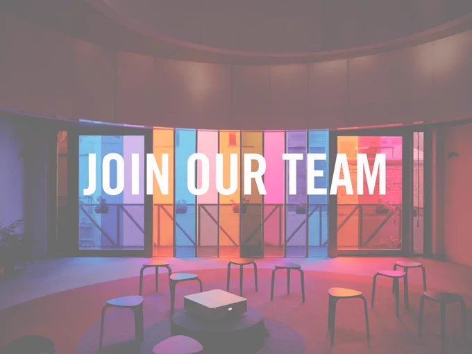 Ten stools stand in an empty room. Glass walls surround flooding the room in hues of orange, blue, pink and purple. The carpet repeats these colours through a circular pattern. Centred large white text reads 'Join our team'.