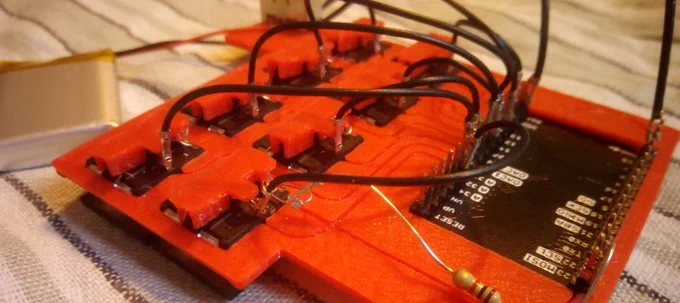 Schwarze Kabel verbinden elektronische Komponenten, die in einem roten Kunststoffrahmen stecken. Die Kabel führen zu Stiftleisten auf einer schwarzen Platine mit weißer Beschriftung. Eine Verbindung besteht aus einem Widerstand statt einem schwarzen Kabel. Ein Lithium-Ionen-Akku ist mit einem roten und schwarzen Draht zu einem Schiebeschalter auf dem Kunsstoffrahmen verbunden. Die Komponenten liegen auf einem grau, schwarz, weiß gewebten Stoff.