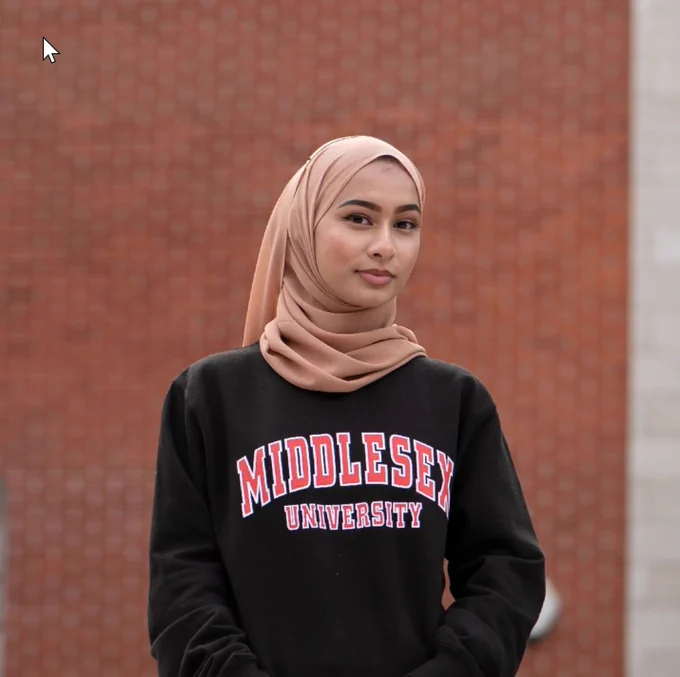 A student wearing a headscarf posing confidently for the camera in from of a red brick wall on campus