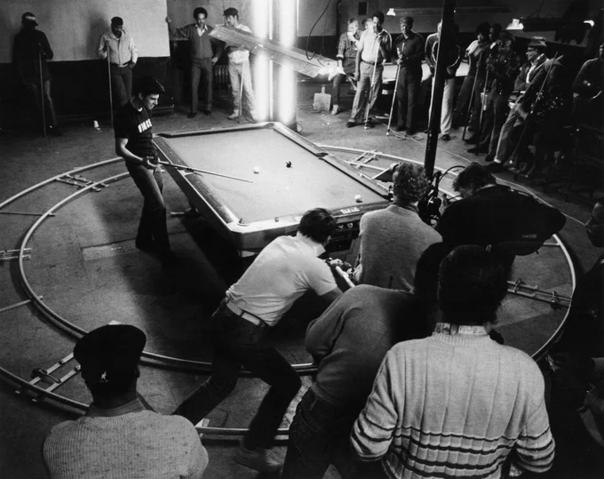 Behind the scenes of The Color of Money. Tom Cruise is at the pool table, cue in hand. Around him and the table is a circular dolly track, cast and crew stand in the edges of the frame, including a camera operator with his kit.