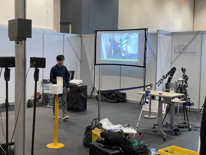 A demo booth with a person setting up a projector. A robotic arm next to that person in front of a table.