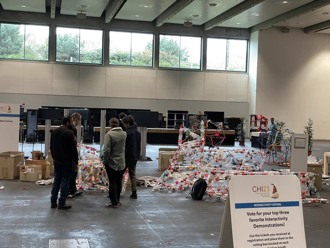 A group of people standing in an exhibition hall, besides a pile of empty bottles that get assembled to a sculpture.