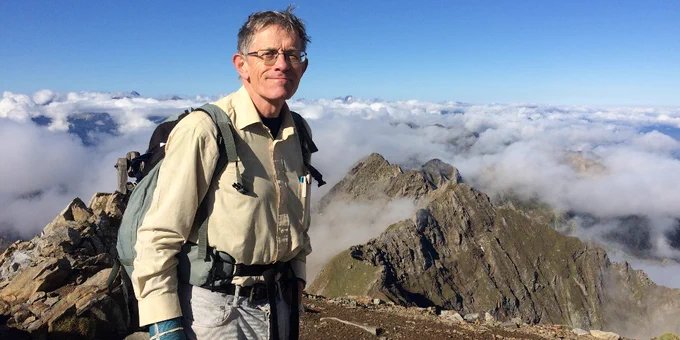 Travel expert standing at the top of a mountain range, in front of a blue sky, cloud inversion and on top of a mountain, looking into the camera