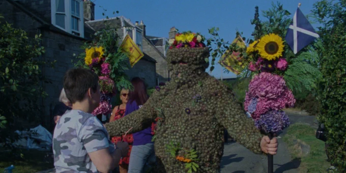 A person dressed head-to-toe in sticky burrs stands with a Scottish flag.