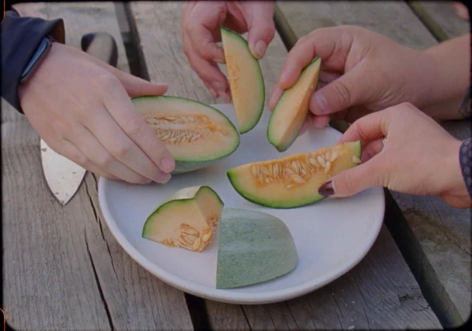 Four hands reach for wedges of melon on a plate.
