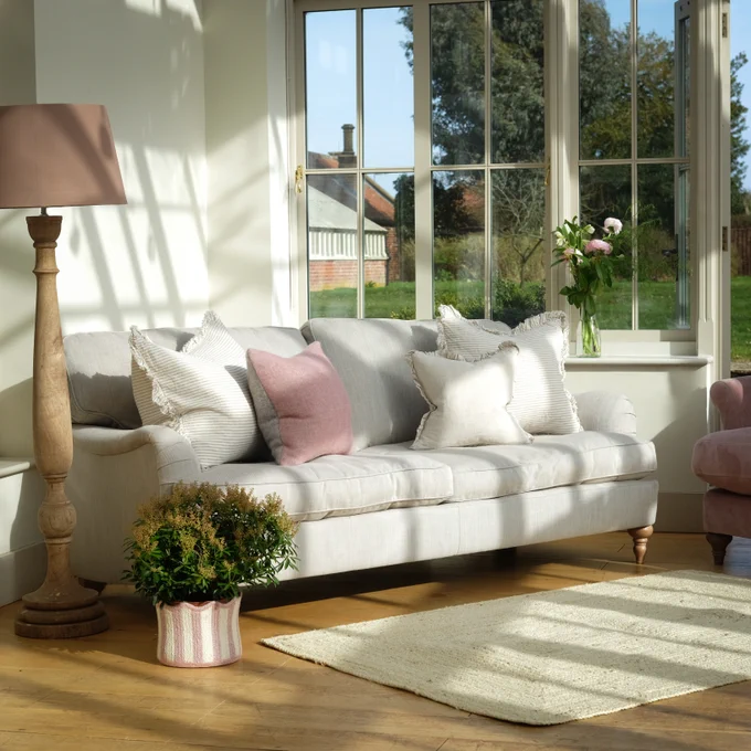 A light grey sofa with white and pink cushions on and a jute matt in front. A tall standing lamp and a short plant stand to the left side, and large windows looking across a grassy garden are behind. 