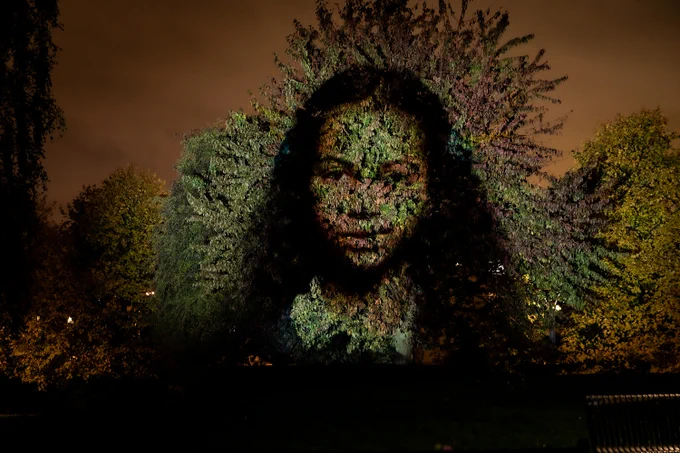 A person's face is projected onto a tree at night.