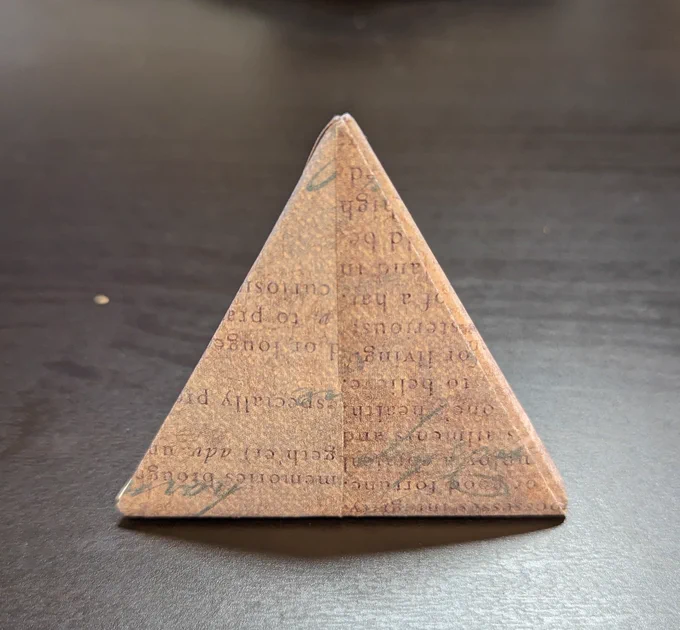 Photo of Origami tetrahedron made of purple paper with abstract blocks of text, from the front. 