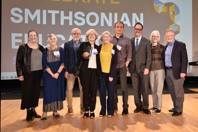 A group of nine adults in business attire pose on stage. A slide on screen behind them reads, Smithsonian Education.