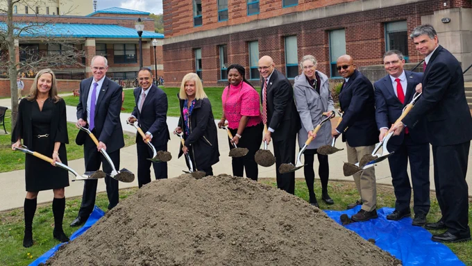 A group of people in business attire hold shovels around a mound of dirt.