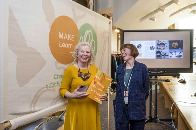 Two figures laugh in front of the MAKE Learn banner and a screen showing craft resources, holding a copy of the MAKE Learn report.