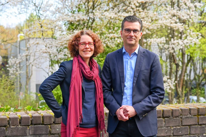 Eine Frau und ein Mann stehen vor einer Mauer, im Hintergrund blühende Bäume.