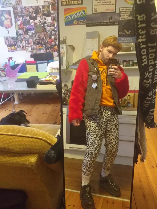 Timi macht ein Spiegelselfie von sich. Sier trägt eine Jacke mit roten Plusch,.dazu einen orangen Pullover mit seiner ihrer Leohose. Der Hintergrund ist eine Fotowand.
