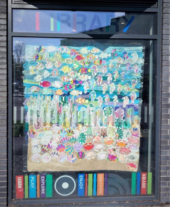 Collage of coloured-in shells, crabs, tropical fish, sea horses, corals and seaweed on a multi-coloured background made to resemble a sandy ocean floor and ocean water, on display in a large window with the word 'LIBRARY' at the top.