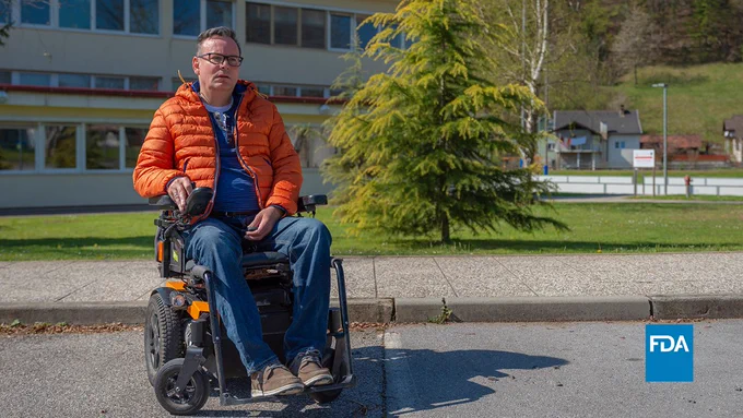 Graphic of a man in a wheelchair wearing jeans and an orange jacket. He is outside in a parking lot with a tree and a building behind him. Blue FDA logo in the bottom right corner