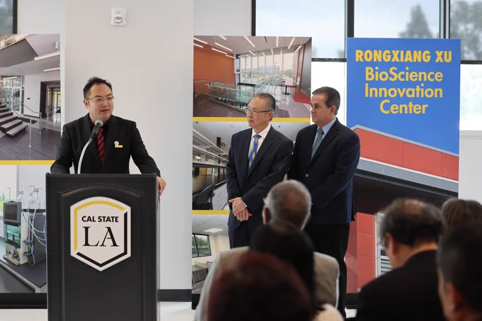 Person speaks at lectern that has Cal State LA badge on it while two others stand by and watch. Banner in the background shows images from inside the center. 
