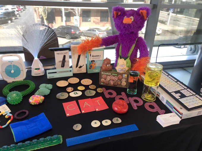 Purple puppet named Edgar along with sensory items (squishy balls, marble maze emotion stones, etc) and literacy tools (CPEN Reader, Scanmarker Reader, Livescribe, tactile letters, etc..) on a table at AT-TIES Together event in Oregon
