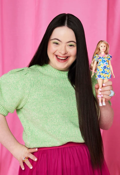 The image shows a white girl with Down's syndrome smiling with her hand on her hip and holding a Barbie doll with Down's syndrome in the other hand. The girl has long dark straight hair and is wearing a dark pink skirt and a pale green jumper. The background is pink