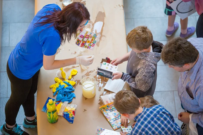 Woman wearing a blue shirt leaning over a table covered in craft paper to put a sticker in a child's sticker book at Science Rendezvous in 2016.