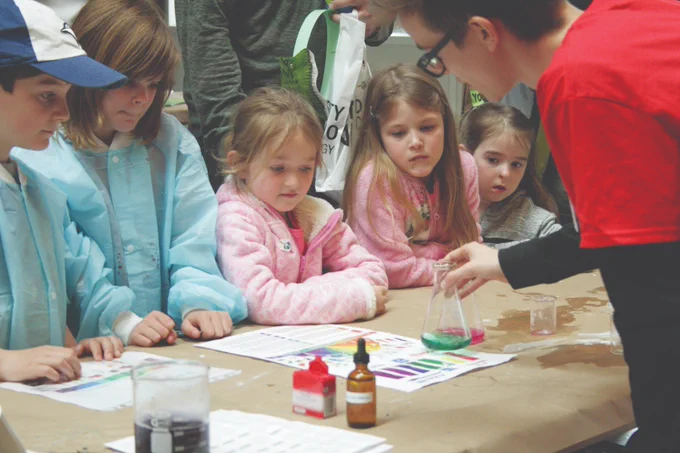 Five children standing behind a table covered with craft paper watching a person in a red shirt hold a beaker with green liquid in it during an experiment at Science Rendezvous in 2017. 